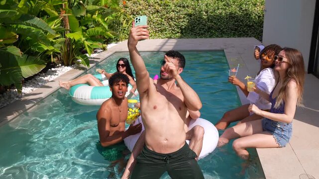 Happy young man taking a group selfie with diverse friends at a swimming pool party, capturing fun summer vacation memories with a smartphone while dancing and drinking cocktails