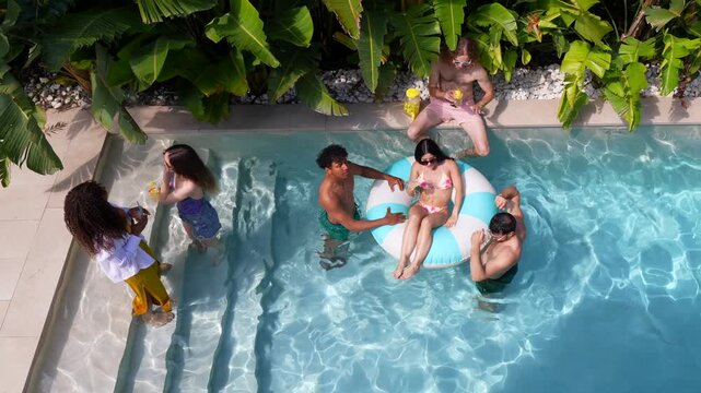 Multiracial group of young friends having fun and drinking cocktails at a summer pool party, with a woman relaxing on an inflatable ring surrounded by lush tropical plants and clear blue water