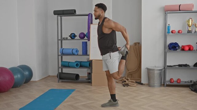 Man in studio holds ankle for standing quad stretch beside exercise mat and shelving with ropes and dumbbells; determination fitness.