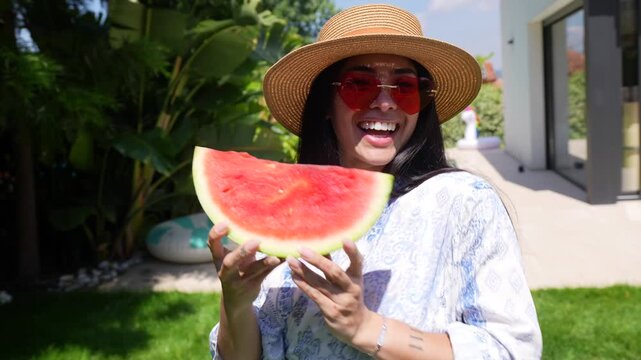 Smiling young woman wearing a straw hat and heart shaped sunglasses eating a fresh slice of watermelon in the backyard garden, enjoying a sunny day during her summer vacation