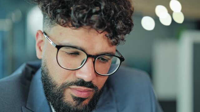 Close up portrait of a pensive male entrepreneur with beard and eyeglasses holding a pen near his chin and contemplating a business strategy. Blurred corporate office interior on background.