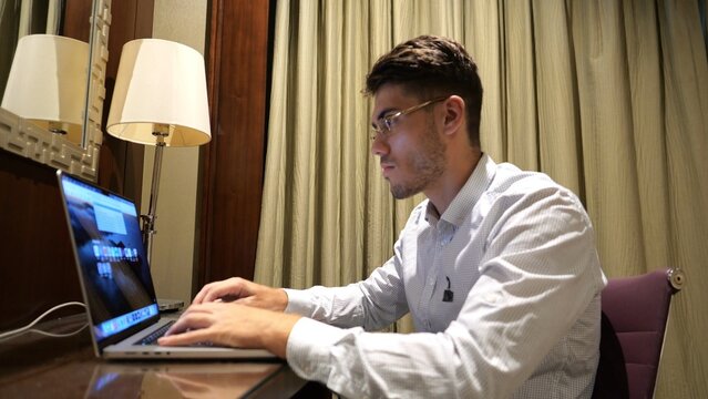 Young businessman in glasses working on laptop in modern hotel room. Handsome entrepreneur using notebook for work at table in home interior. Manager showing remote job and productivity. Close up