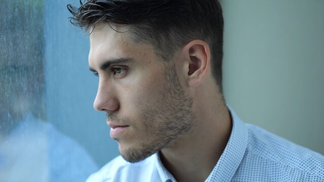 Portrait of sad young entrepreneur sitting near window in modern hotel room or office. Unhappy man with serious expression looking away showing emotional stress, fatigue and professional burnout