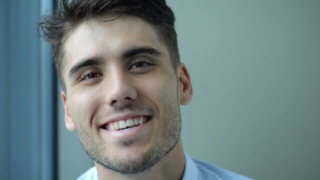 Portrait of happy businessman sitting near window in modern hotel room or office. Relaxed young manager looks into camera with smile showing relief, confidence and success in professional lifestyle