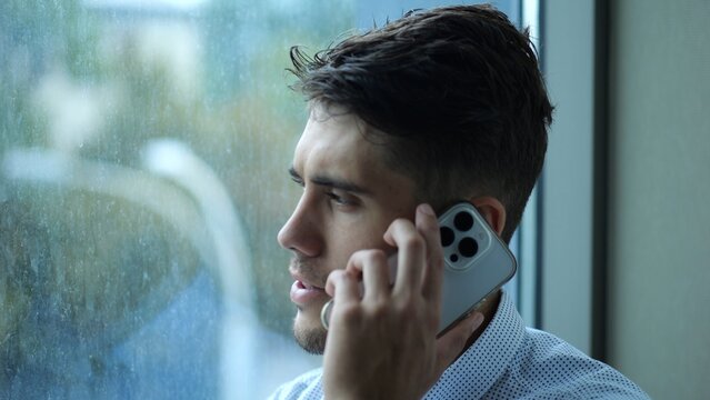 Portrait of male entrepreneur in shirt speaking by smartphone near large window into skyscraper. Young confident businessman talk on phone at office. Handsome manager having conversation on cellphone
