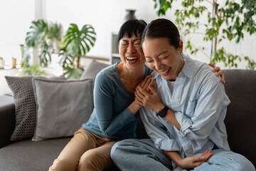 Fototapeta premium Two women laughing together on sofa at home, sharing joyful emotions, friendship and warm companionship in a cozy modern living room