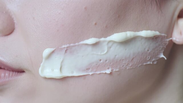 Asian female applying cream to her cheek with a finger, showcasing a smooth texture and gradual spreading of the product across her skin in a close-up shot