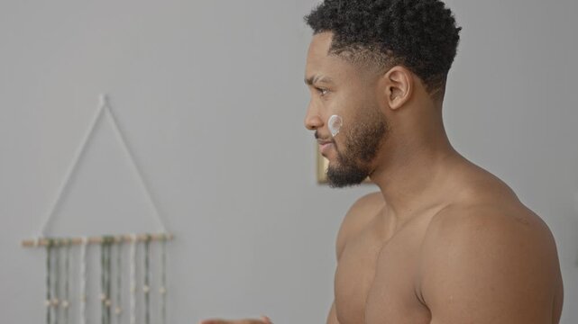 Shirtless man presses hands to face while applying cream to cheek in building beside mirror and macrame wall hanging; introspection calm.