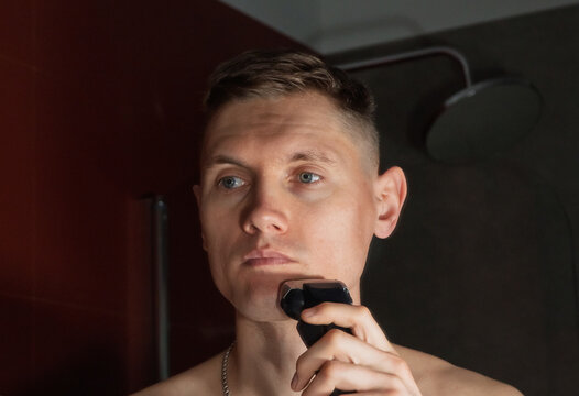 Attractive European-looking man shaves with an electric razor in front of a bathroom mirror. Young man using an electric razor while shaving in a modern bathroom during his morning grooming routine. 