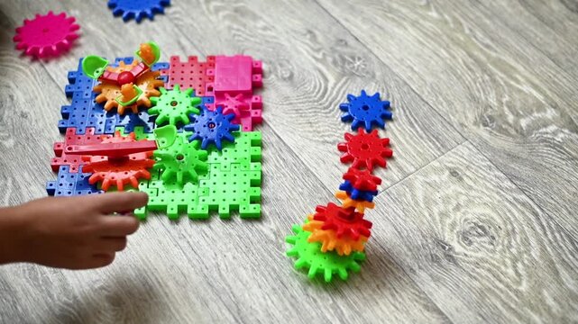 Constructor toy with colorful plastic gears arranged on floor while child hand builds mechanism. Educational constructor encourages creativity, motor skills, logical thinking