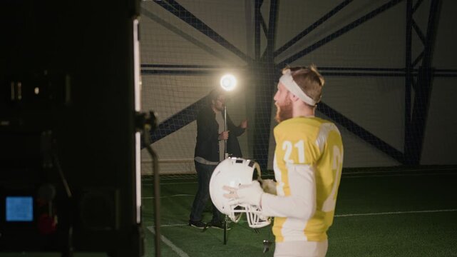 Medium side backstage shot of young Caucasian male American football player in yellow jersey, protective armor standing on pitch, posing for commercial shoot while gaffer is setting up lights