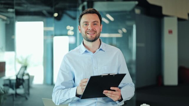 Male entrepreneur or manager CEO writing on a clipboard, looking up at camera with a confident smile. Happy professional corporate worker holding document report inside modern office interior.