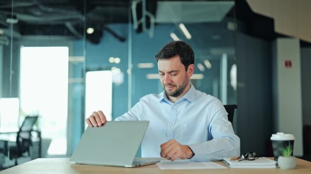 Male manager closing laptop computer, leaning back in office chair with hands behind head, smiling and celebrating project completion. Pleased corporate worker enjoying successful end of workday.