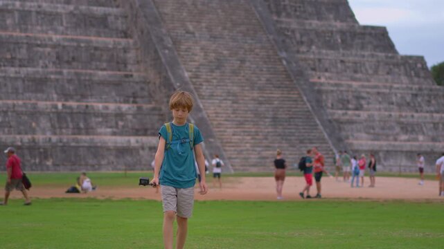 A boy recording himself on action camera while walking in front of Kukulkan pyramid in Chichen Itza, Mexico. Travel vlog, childhood adventure and tourism content creator concept. Slowmotion video.