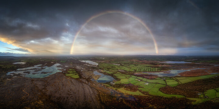 Aerial view of a vibrant full rainbow arching over the limestone pavement and green fields of the Burren under dramatic storm clouds in Burren, County Clare, Ireland.