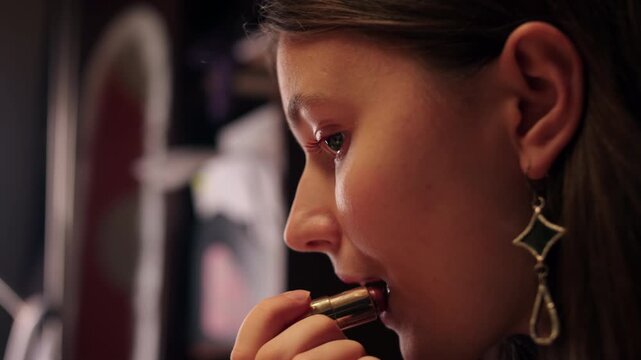 Lipstick application scene when young caucasian woman beautifying before evening event. Profile shot of brunette woman applying makeup in a room with dim light. Red lipstick is applied to female lips