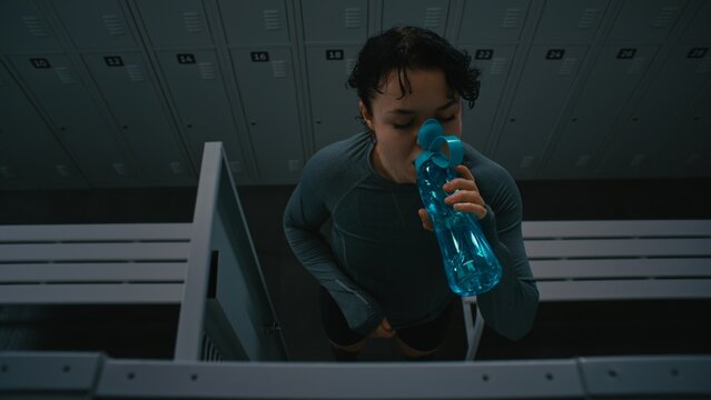 Fit Woman in Locker Room Puts on Performance Long Sleeve Over Sports Bra and Shorts. Standing Between Benches and Lockers, Female Finishes Preparation by Opening Water Bottle. Healthy Lifestyle.