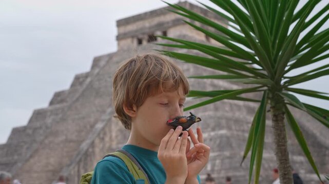 Little boy blowing bird shaped whistle in front of Kukulkan pyramid in Chichen Itza, Mexico. Child playing traditional toy at famous landmark. Video with sound.