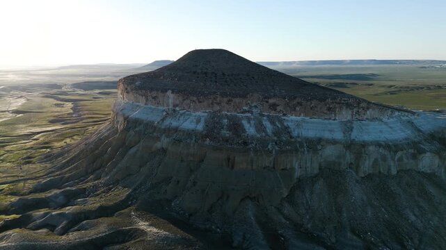 Drone view of the shaded side of Mount Sherkala on a warm summer evening. Famous landmark of Mangystau surrounded by green grass under a clear sky. Unique geology of Kazakhstan, limestone cliffs, 