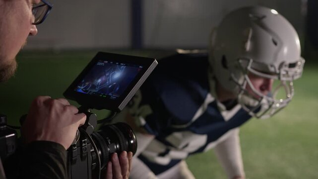 Medium close-up of cameraman recording video of American football player in protective helmet crouching in powerful stance for scrimmage with rival team while training at stadium
