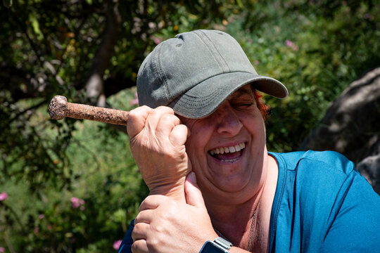 Una mujer sonriente con gorra gris y camiseta azul sostiene un tornillo junto a su cabeza en un gesto divertido, rodeada de &aacute;rboles y flores bajo la luz del sol.