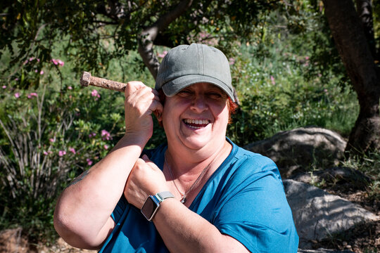 Una mujer sonriente con gorra gris y camiseta azul sostiene un tornillo junto a su cabeza en un gesto divertido, rodeada de &aacute;rboles y flores bajo la luz del sol.