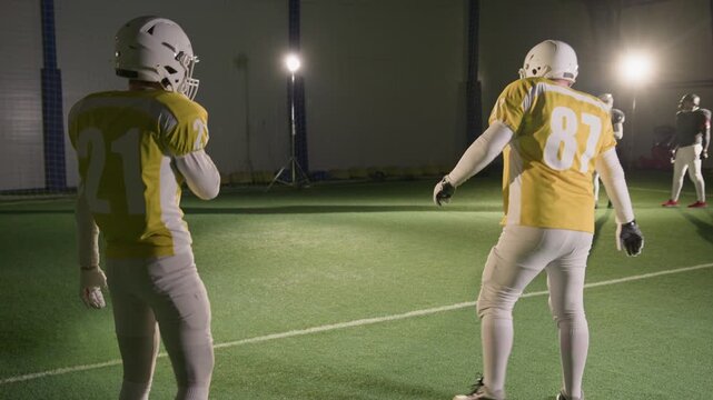 Wide tilt-up shot of team of American football athletes practicing kickoff and ball catch while training at stadium, in full uniform and protective gear, filmed by cameraman for reality sports show