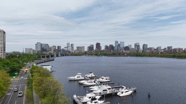 Aerial drone view looking down the Charles River in Boston, featuring luxury boats docked at a marina with the historic Longfellow Bridge, Storrow Drive traffic, and city skyline in the background.