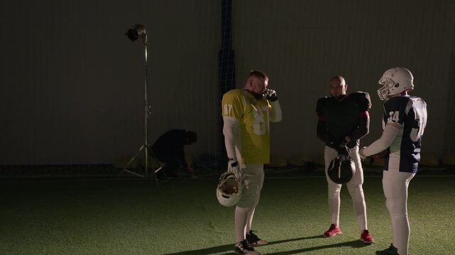 Wide shot of three American football team players standing together at arena before training session, talking while male gaffer is setting up lights to shoot commercial or reality series content