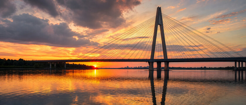 Serene sunset over a calm lake with a cable-stayed bridge silhouette, reflecting warm orange and golden light across the water in a peaceful, cinematic sky.