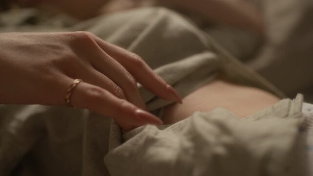 Close up shot of hand of unrecognizable woman with long nails unbuttoning linen shirt and revealing part of back while relaxing in bed in morning