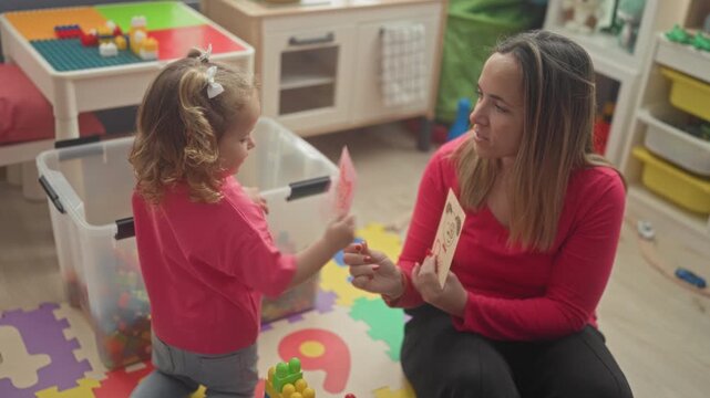 Woman holds a flashcard while little girl holds a pink card to her face amid foam mats and toys in a playroom building; focused early learning.