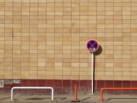 Yellow tiled wall with red base and white no parking sign with blue symbol next to orange barriers and white metal railings in urban setting.