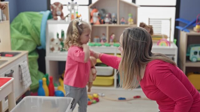 Woman handing doll to toddler while seated on foam playmat in studio with storage shelves, toy bin and scattered blocks visible; nurturing playful bonding.