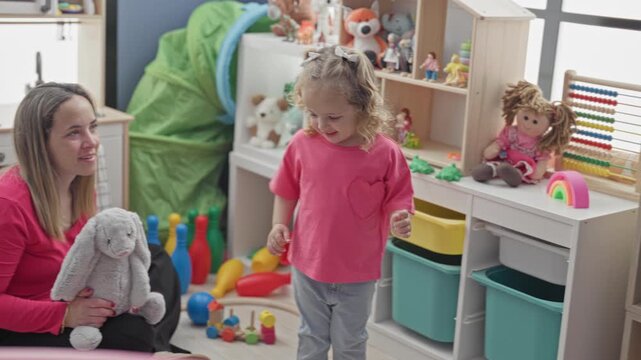 Toddler girl reaches hand toward toy bin by a chalkboard while seated woman guides and watches in a bright playroom; nurturing play.