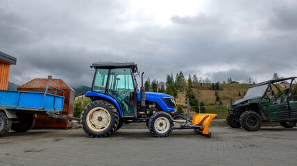 Obraz premium Blue tractor with snow plow attachment and trailer parked next to ATV