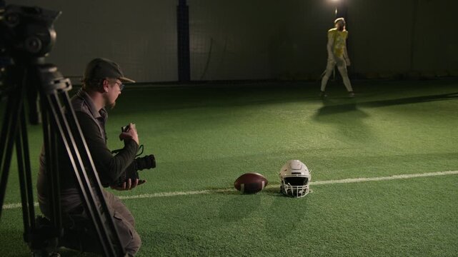 Full arc shot of professional male videographer kneeling on turf on pitch, filming gridiron football helmet and ball from ground level for documentary b-roll while team warming up for training