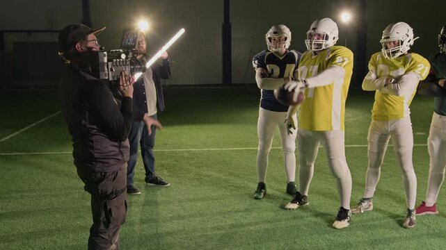 Full backstage shot of cameraman filming portrait of Caucasian male American football team dressed in yellow team jerseys, helmets, protective armor for club promotion, gaffer holding light stick