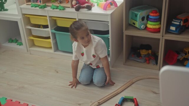 Hispanic girl kneels and reaches arranging toy cars on a wooden train track beside foam alphabet mat and storage shelves inside a playroom building; concentration imagination play.