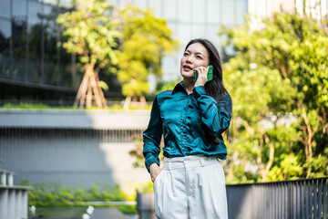 Fototapeta premium Confident young Asian businesswoman talking on smartphone in outdoor urban park setting. Smiling female professional wearing elegant teal shirt communicating on mobile phone outside office building.