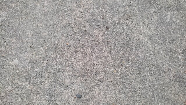 This overhead close up photograph shows a weathered grey concrete surface texture with small pebbles and rough grit distributed across the entire flat ground plane area