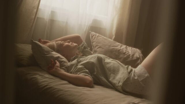 Handheld shot of female Caucasian zoomer lounging and lazily stretching after waking up under morning sunlight in bed