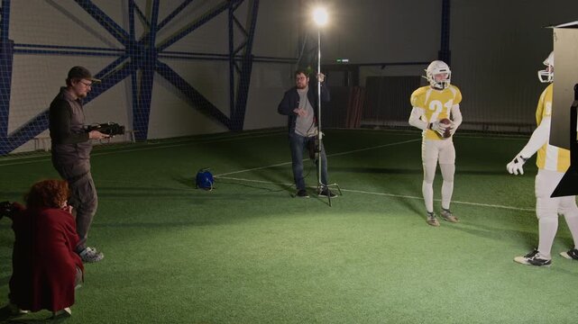 Wide backstage shot of Caucasian male filmmaker directing gridiron football players to film victory scene, adjusting video camera, producer and gaffer observing, working together on sports show set