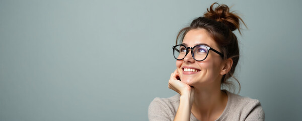 Fototapeta na wymiar Young woman with glasses smiles, looking up thoughtfully. She rests chin on hand, thinking about new ideas. Casual indoor studio portrait with copy space.