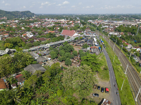 Aerial view of the Animal Market Prambanan with livestock under trees near railway tracks and urban rooftops in Kecamatan Prambanan, Jawa Tengah, Indonesia.