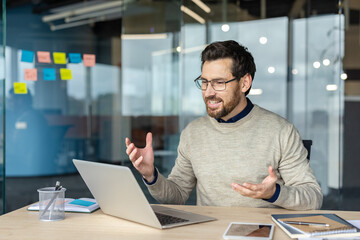 Fototapeta premium Bearded man wearing glasses engaging in a video call and gesturing while communicating on a laptop from his modern office workspace, showcasing remote work and online connection