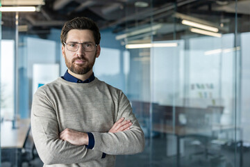 Obraz premium Confident bearded businessman wearing eyeglasses and a casual sweater, standing with crossed arms in a contemporary corporate office, portraying professionalism and leadership