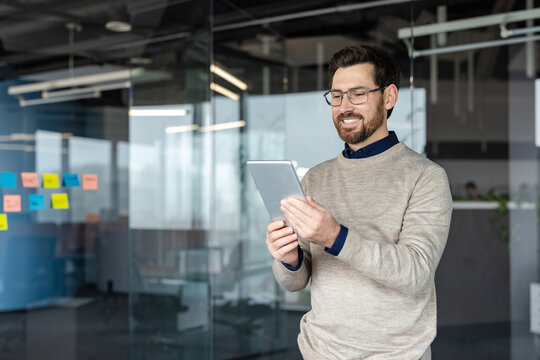 Smiling man standing in a modern office, wearing glasses and a sweater, holding a digital tablet and working while looking at the screen with a happy expression