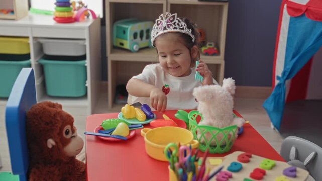 Hispanic girl wearing tiara holds a plastic spoon with her right hand and serves toy fruit to a teddy on a red kids table inside a building; playful imagination.