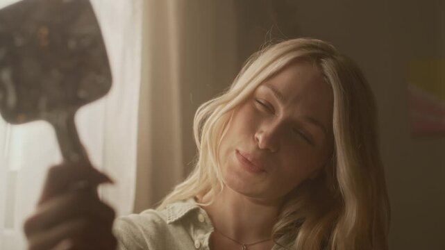 Close up shot of blonde gen Z Caucasian woman looking at herself in mirror reflecting sunlight patterns while relaxing during morning home weekend